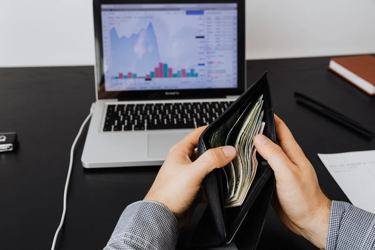 Man holding a leather wallet with bills in front of a laptop displaying financial graphs.