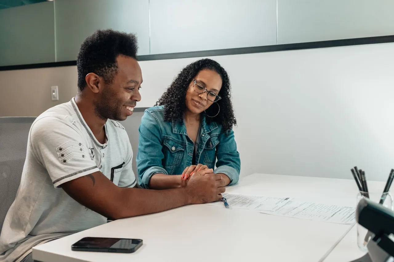 A couple sits at a white table, happily reviewing documents and holding hands in an office setting.